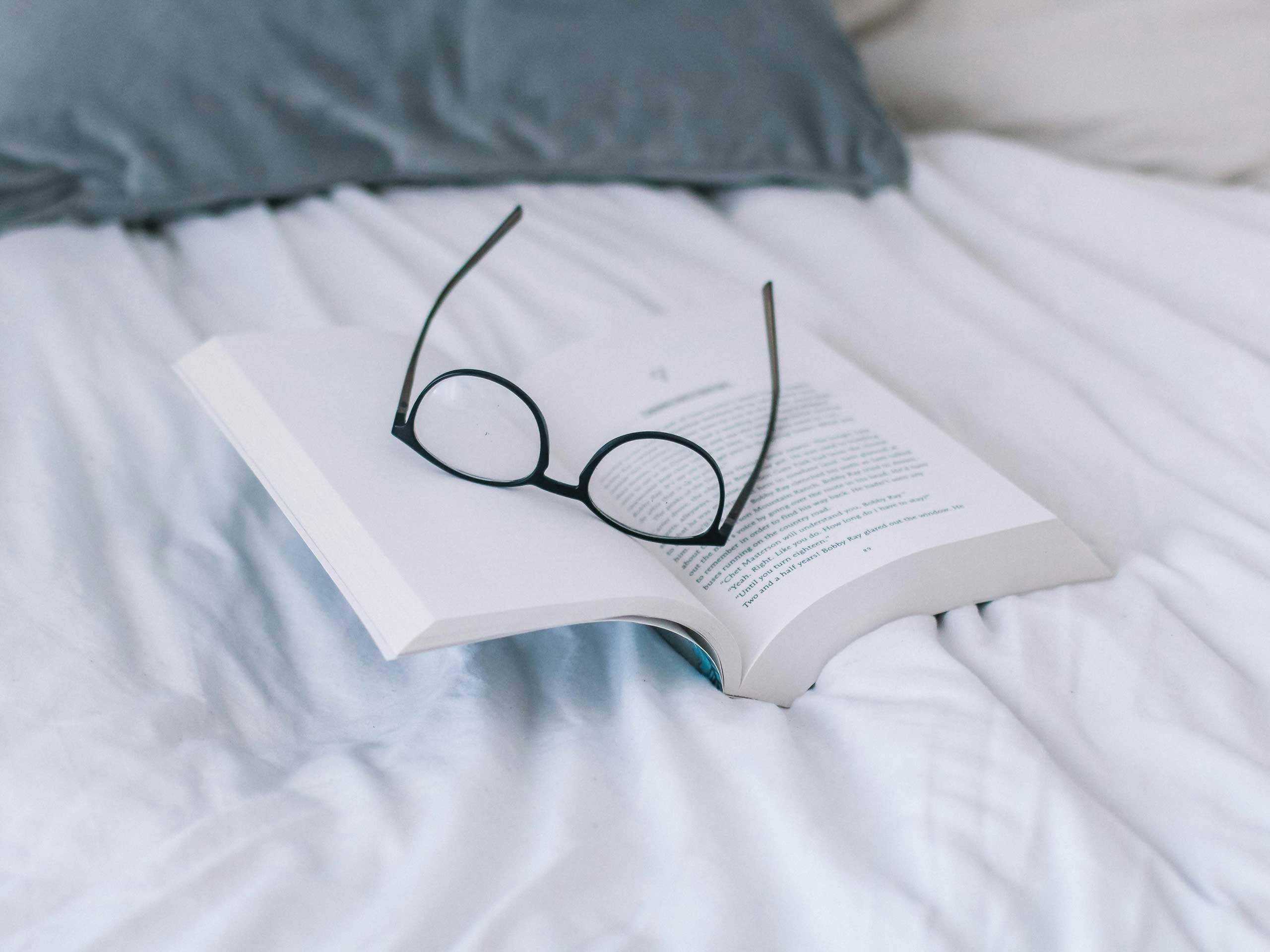 Aufgeschlagenes Buch mit Brille auf einem hellen Bett.
