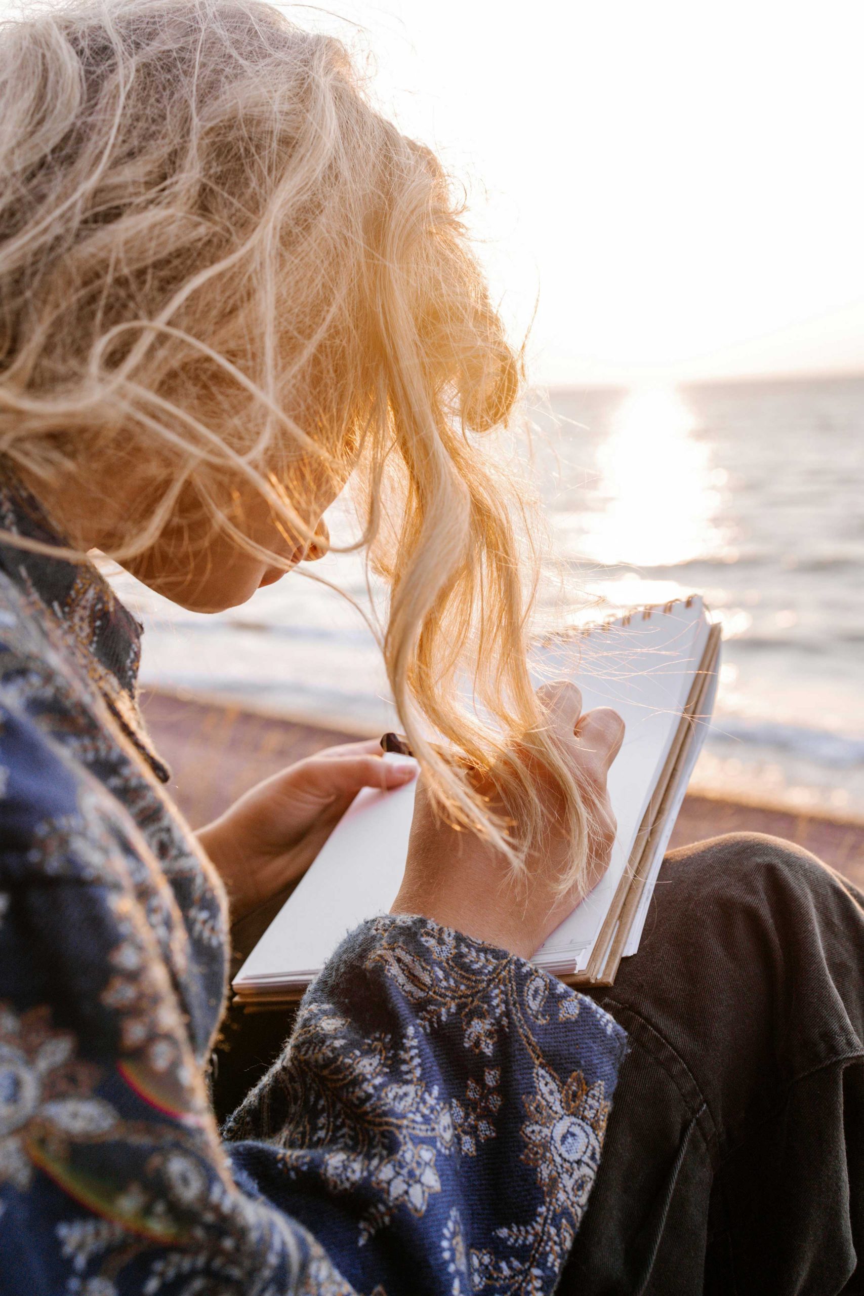 Person mit langen blonden Haaren sitzt am Strand im Sonnenlicht.